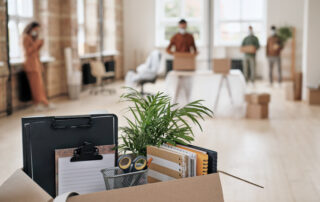 Focus on open cardboard box with office supplies, clipboards and plant against people moving into new office in background